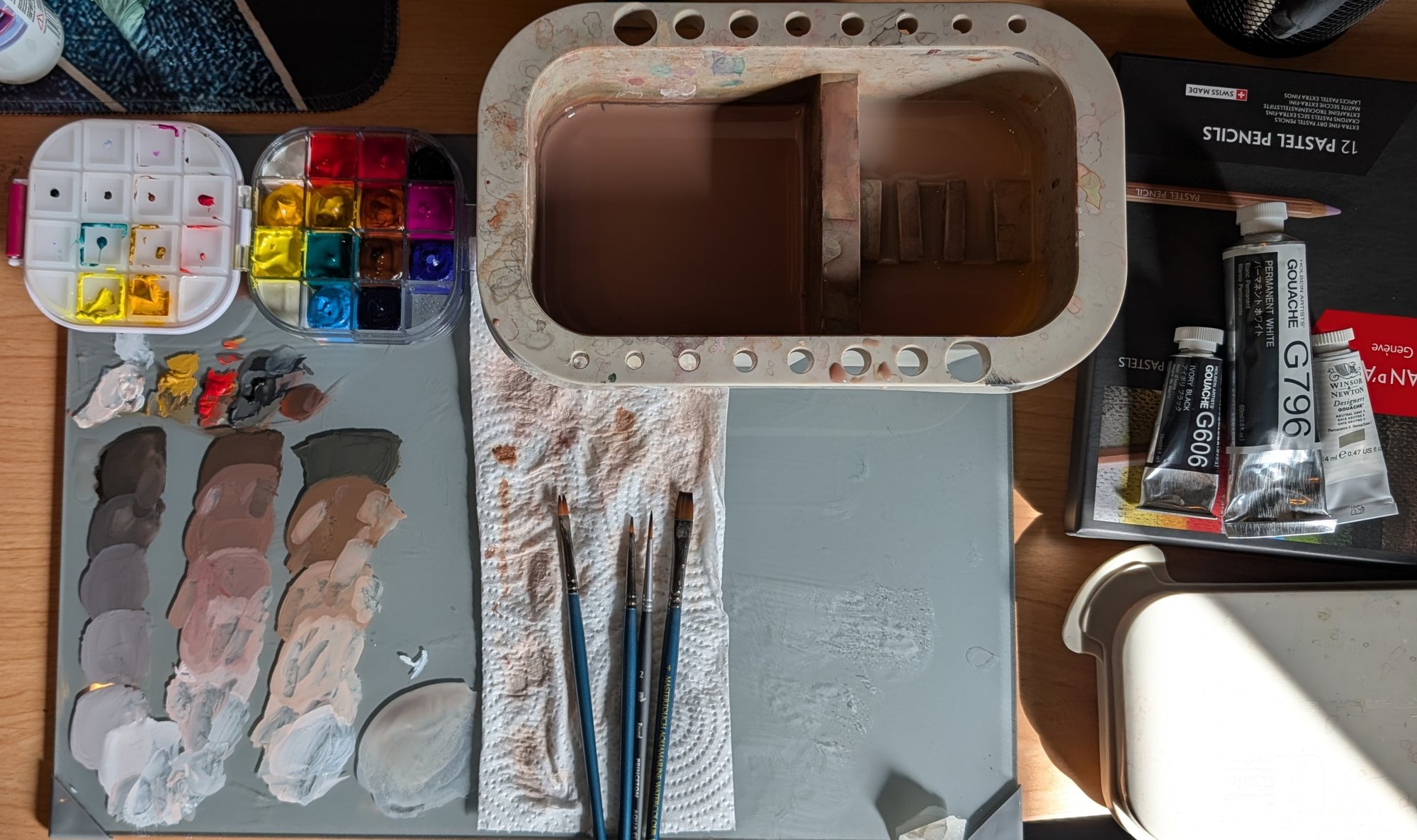 The working setup after the last head: a grey mixing palette with three value strings, brushes, a water pot, gouache tubes, a black pastel pencil, and an airtight paint palette at top left